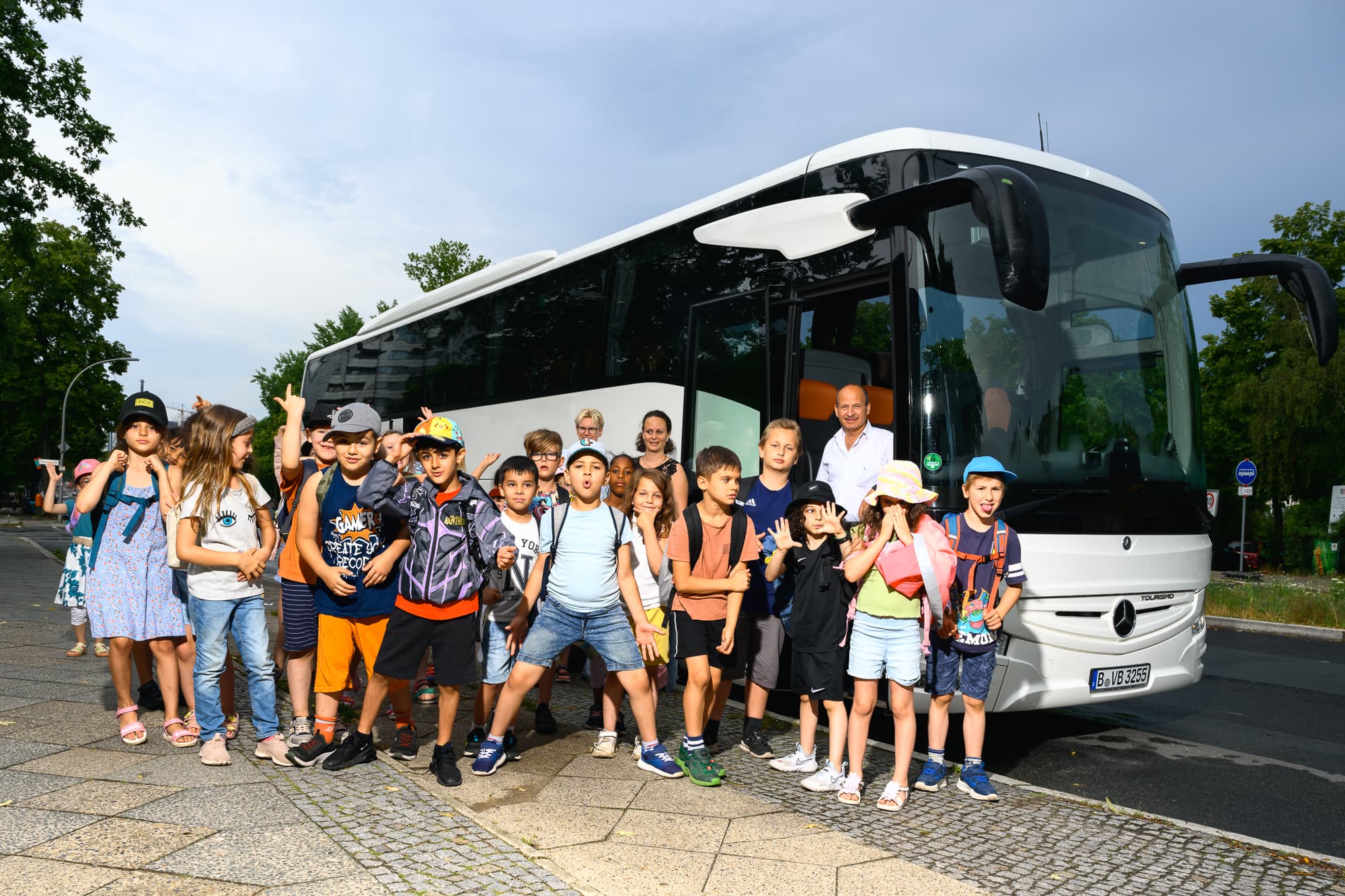 Eine Kindergruppe mit Kindern im Grundschulalter stehen vor einem weissen Bus für ein Gruppenfoto zusammen. Ein Junge im Vordergrund macht eine coole Pose im Hiphop-Style.